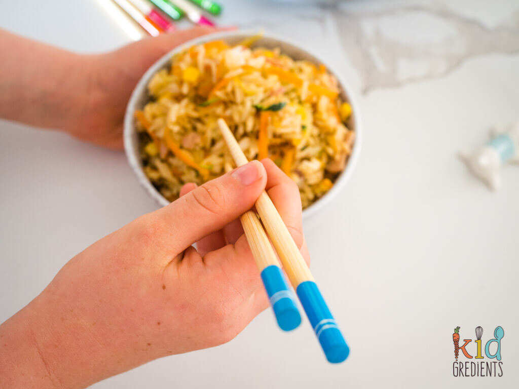 child's hands holding blue handled chopsticks and a bowl of fried rice
